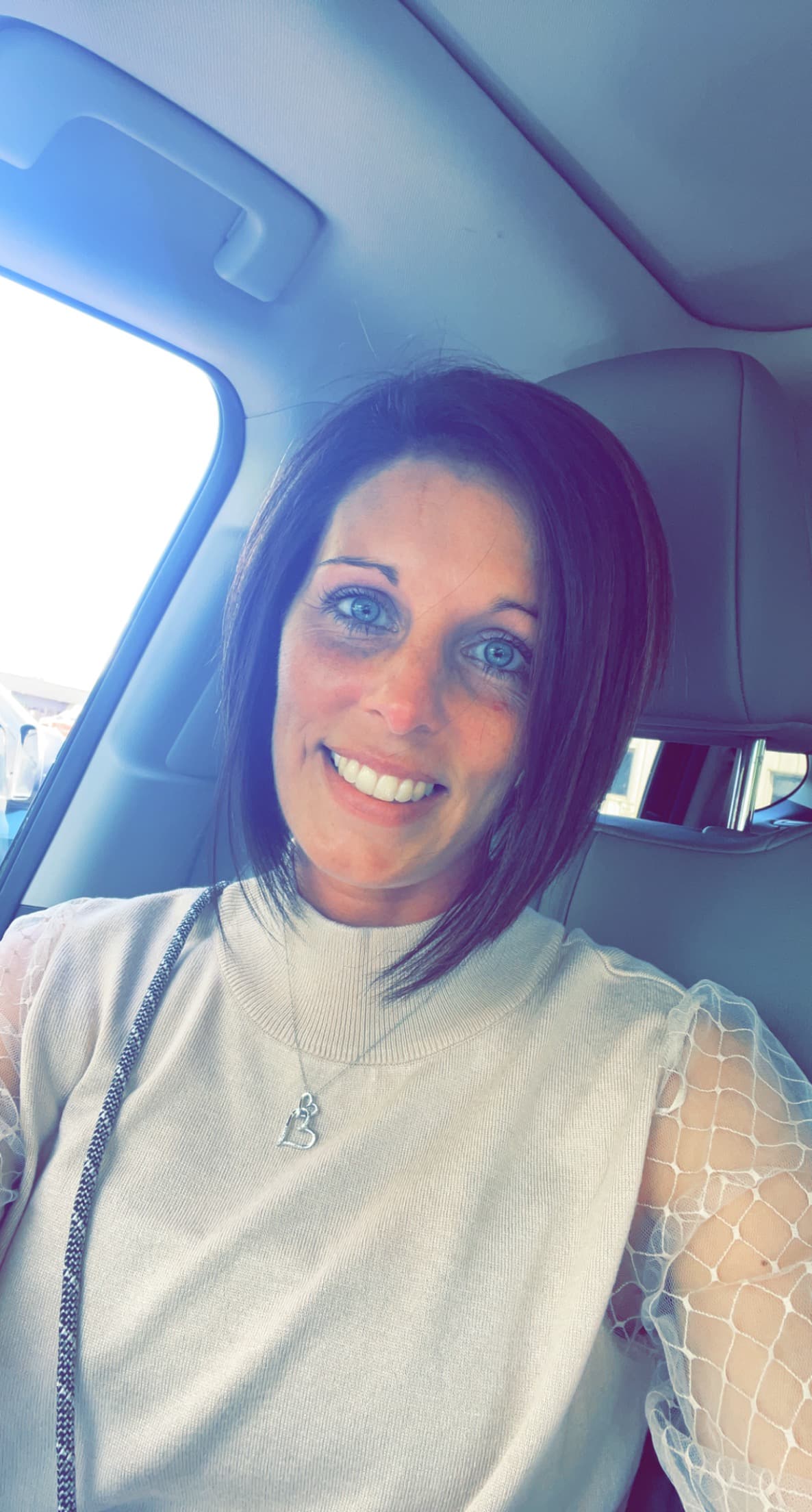 Smiling woman with short brown hair and blue eyes wearing a cream top in car.